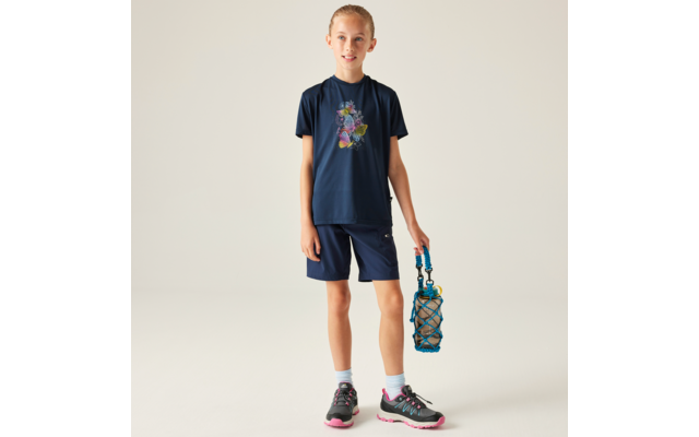Kind steht vor neutralem Hintergrund, trägt dunkelblaues T‑Shirt mit buntem Motiv, dunkelblaue Shorts, Sportschuhe und hält eine blaue Trinkflasche in einem Netz