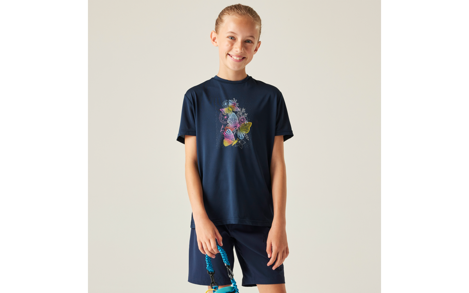 Person in dunkelblauem T‑Shirt mit buntem Schmetterlingsdruck und dunkelblauen Shorts, lächelnd, hält eine blau‑türkise Tasche vor hellem Hintergrund