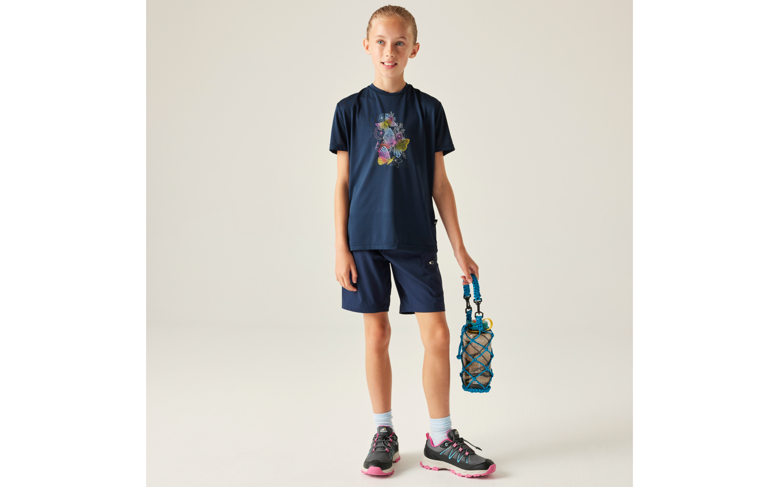 Kind steht vor neutralem Hintergrund, trägt dunkelblaues T‑Shirt mit buntem Motiv, dunkelblaue Shorts, Sportschuhe und hält eine blaue Trinkflasche in einem Netz