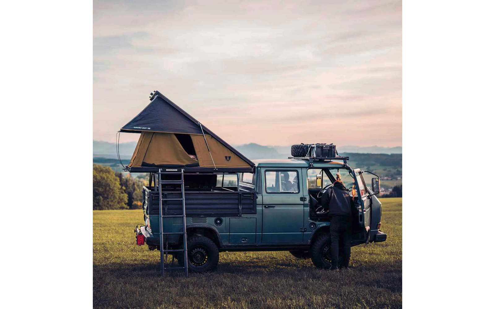 Blaugrünes Pickup-Fahrzeug mit aufgeklapptem Dachzelt, Leiter, Gepäck auf dem Dachträger, eine Person an einer offenen Tür, Wiese, Hügel im Hintergrund, rosa-oranger Himmel