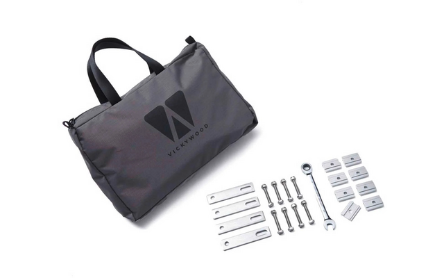 Graue Tasche mit schwarzen Tragegriffen und aufgedrucktem Logo; rechts davon mehrere flache Metallplatten, Schrauben mit Muttern, ein Maulschlüssel und weiße rechteckige Kunststoffteile