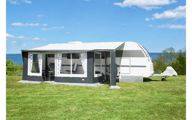Witte caravan met grijs voortent op een groen grasveld, zee op de achtergrond en blauwe lucht met wolken