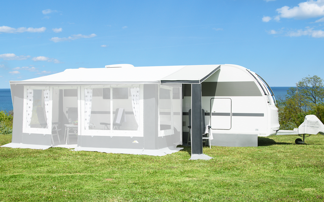 Weißer Wohnwagen mit angebautem Vorzelt auf einer Wiese, blauer Himmel mit wenigen Wolken, Meer und Bäume im Hintergrund
