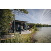 Wohnmobil mit ausgefahrener Markise am Ufer eines Sees, zwei Personen am Wasser, Solarpanele auf dem Gras, Bäume und Himmel mit Kondensstreifen