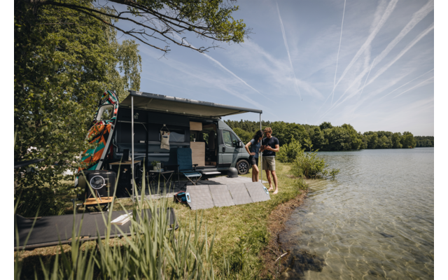 Wohnmobil mit ausgefahrener Markise am Ufer eines Sees, zwei Personen am Wasser, Solarpanele auf dem Gras, Bäume und Himmel mit Kondensstreifen