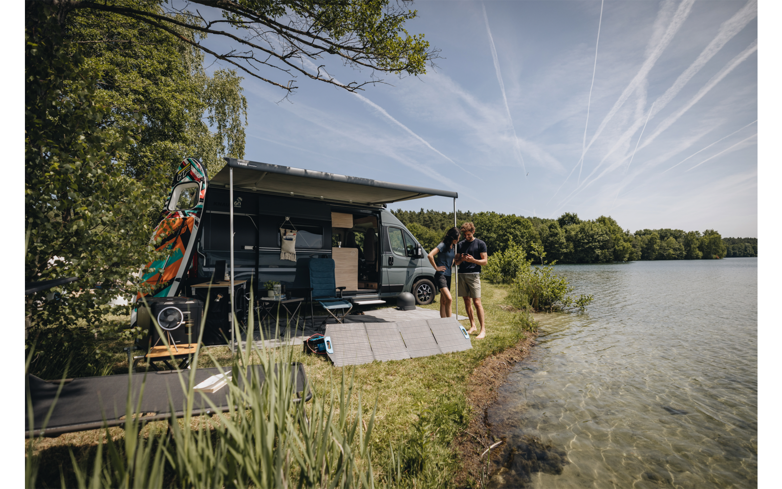 Wohnmobil mit ausgefahrener Markise am Ufer eines Sees, zwei Personen am Wasser, Solarpanele auf dem Gras, Bäume und Himmel mit Kondensstreifen