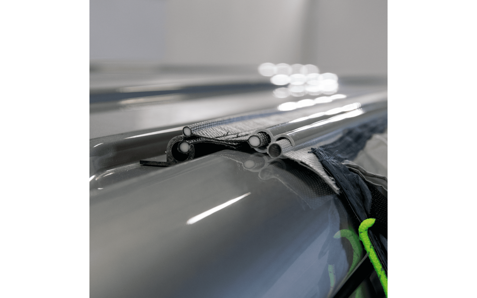 Shiny dark gray curved surface, two parallel silver metal tubes with visible cross-sections, folded dark fabric between the tubes, bright light reflections, green strap at lower right