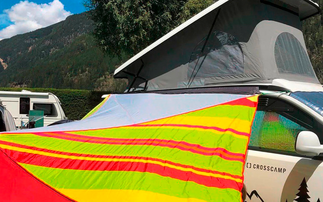 Camper blanc avec toit relevé et auvent rayé jaune-rouge-vert vif fixé dessus, montagnes, arbres et ciel bleu en arrière-plan