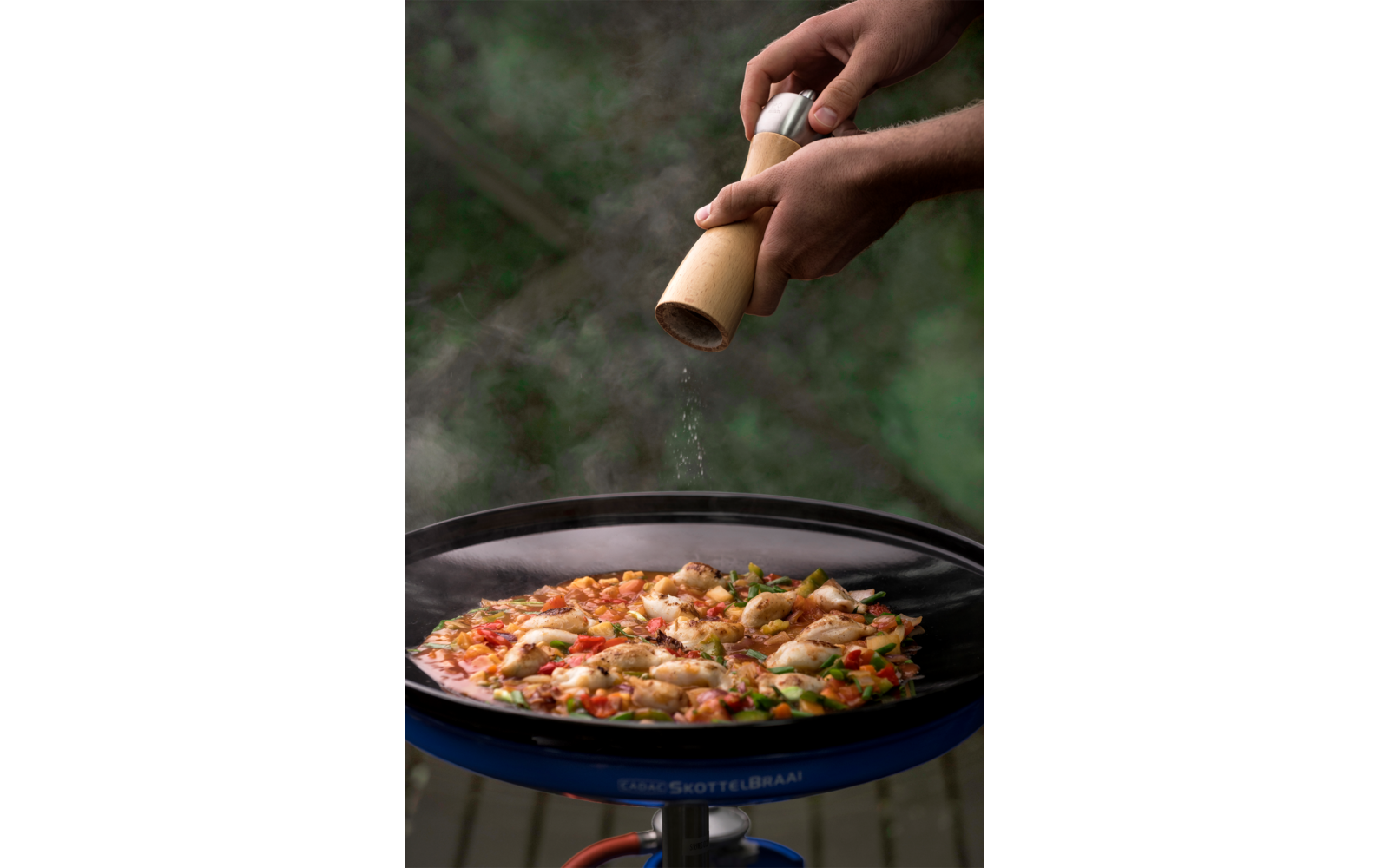 Zwei Hände halten eine Holzgewürzmühle; Partikel fallen auf eine Pfanne mit buntem Gemüse und hellen Stücken; unscharfer grüner Hintergrund