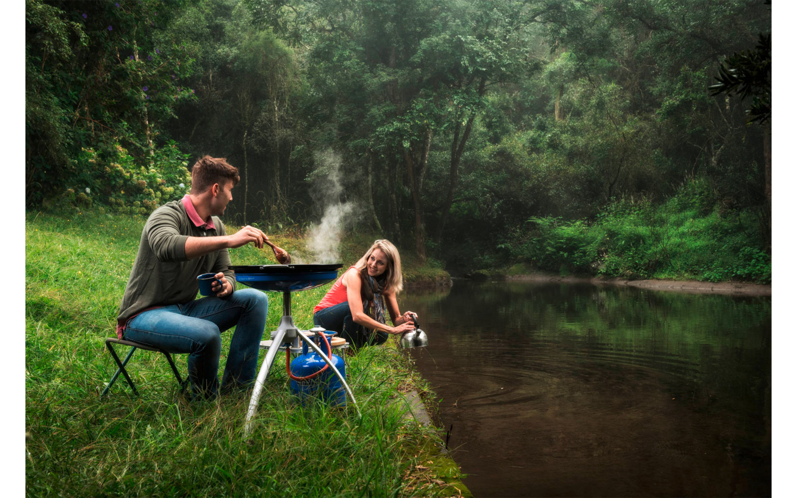 Zwei Personen am Flussufer: eine rührt in einer Pfanne auf einem tragbaren Kocher auf einem kleinen Tisch, die andere kniet am Wasser und hält einen Topf; Grasufer und dichter Wald im Hintergrund