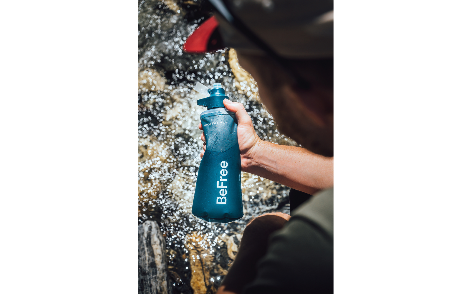 Person hält blaue Trinkflasche mit Aufschrift "BeFree" nahe fließendem Bach; Arm und Hut sichtbar