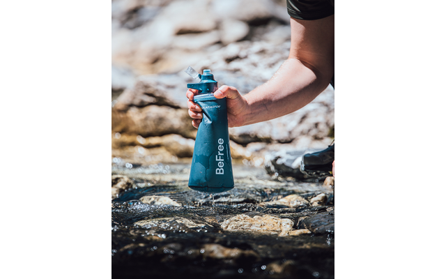 Hand hält eine blaue Wasserflasche mit der Aufschrift 'BeFree' über einem flachen, steinigen Bach