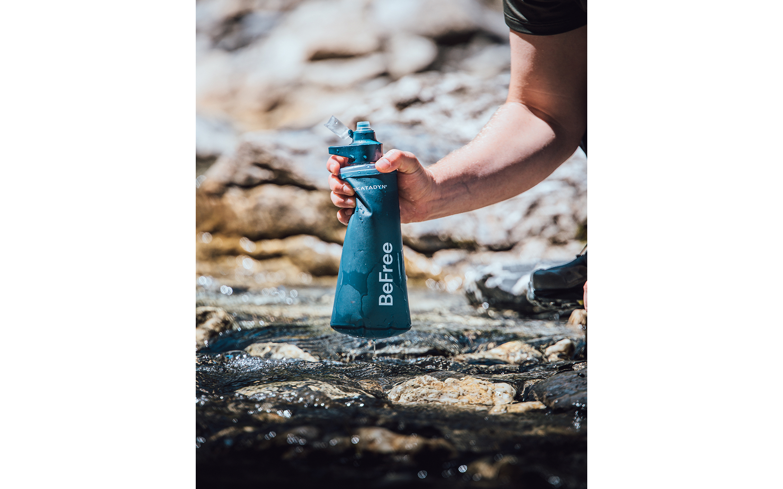 Hand hält eine blaue Wasserflasche mit der Aufschrift 'BeFree' über einem flachen, steinigen Bach