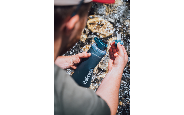 Hände einer Person halten eine dunkelblaue Trinkflasche mit Aufschrift "BeFree" und einen kleinen Filteraufsatz über fließendem Wasser mit sichtbaren Steinen