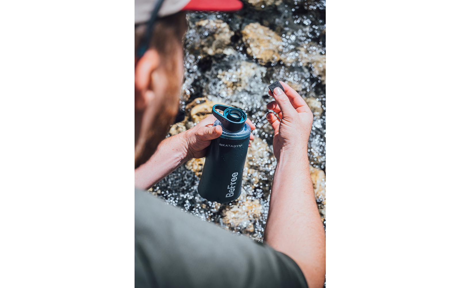 Person von hinten hält eine schwarze Trinkflasche mit abgenommener Kappe über einem flachen, steinigen Bach