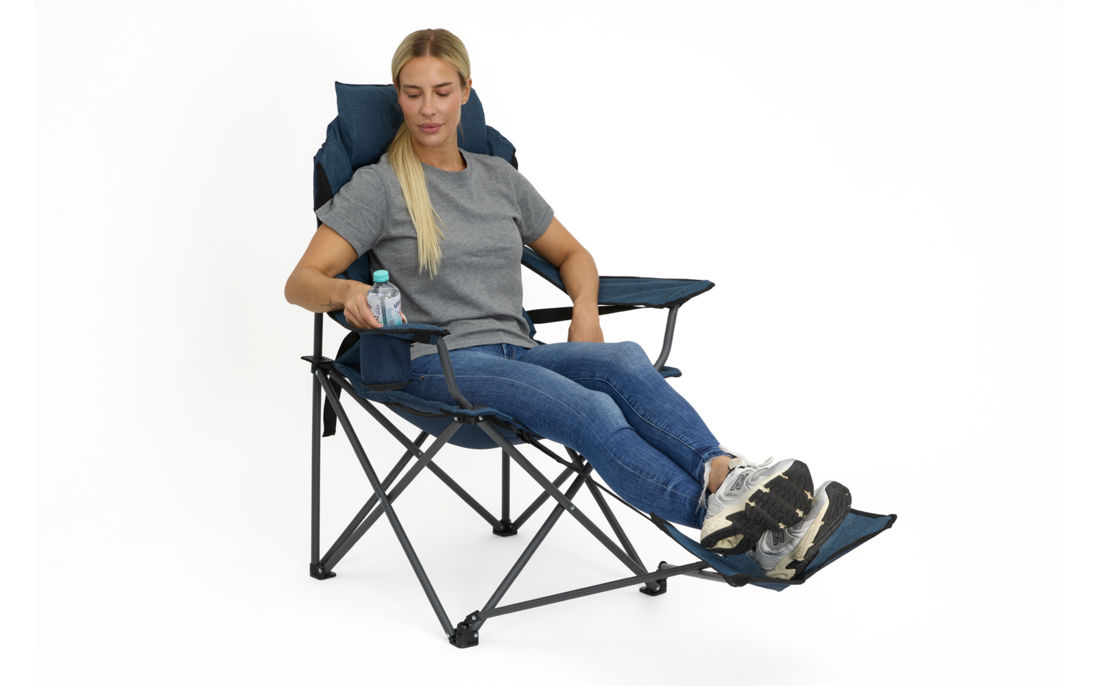 Frau sitzt in blauem Klappstuhl mit ausgeklappter Fußstütze, trägt graues T‑Shirt, Jeans und Sneakers, hält eine Wasserflasche; weißer Hintergrund