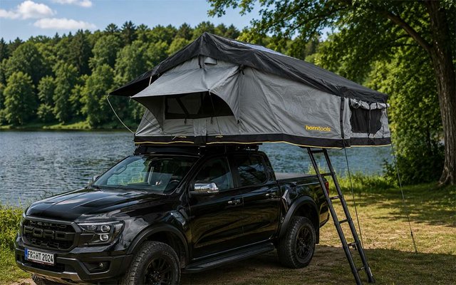 Pick-up noir au bord d’un lac avec une tente de toit déployée et une échelle, forêt et arbres en arrière-plan, herbe au premier plan