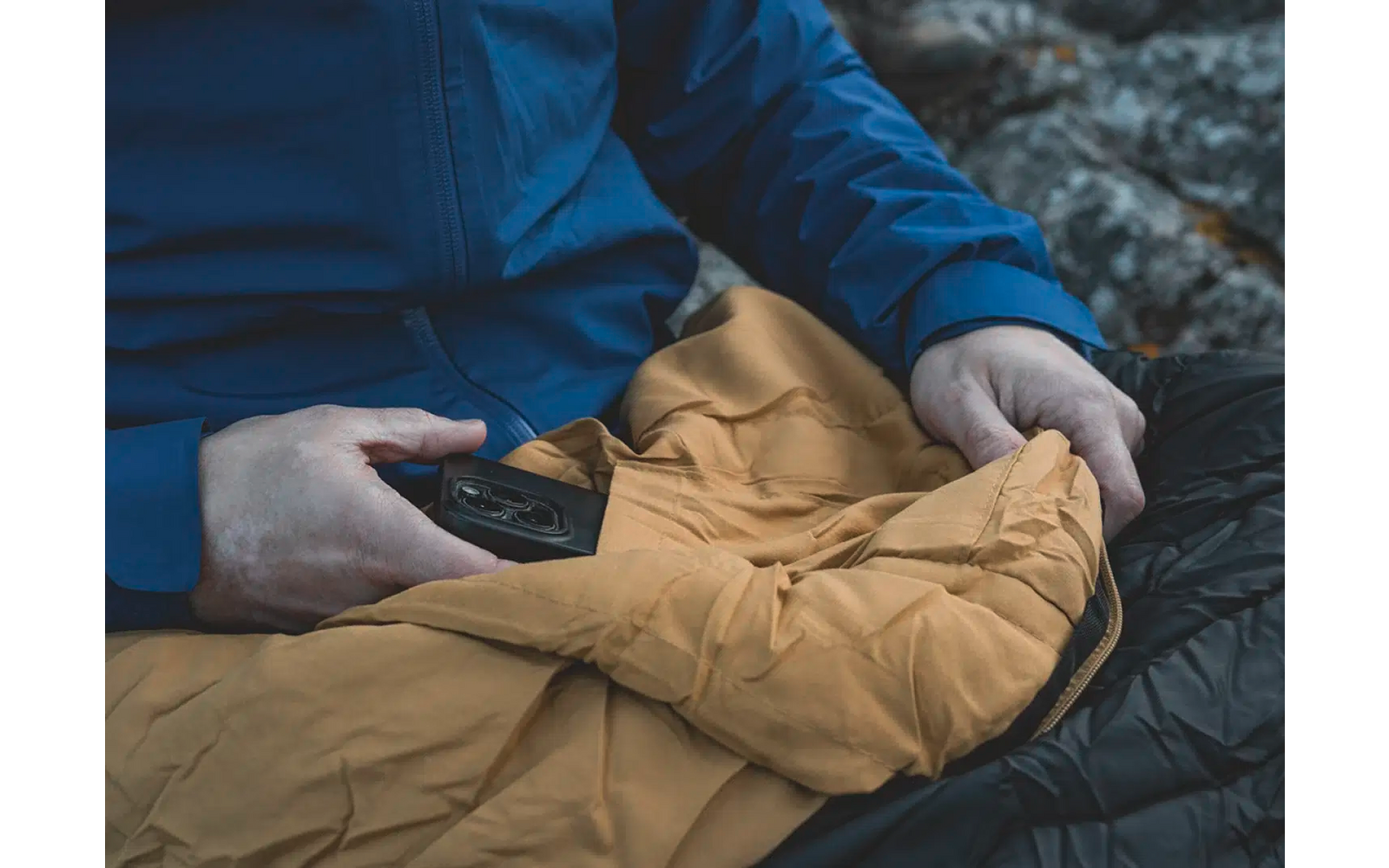 Een persoon houdt een gele jas en een smartphone vast, zittend op donkere rotsen