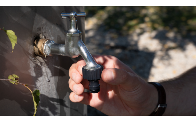 Hand hält einen Metallwasserhahn mit schwarzem Schlauchanschluss, im Hintergrund unscharfe Steine und Pflanzen