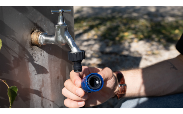 Metallwasserhahn an einer Außenwand, eine Hand hält einen blauen Schlauchanschluss, Unterarm mit Armbanduhr, Boden und Schatten im Hintergrund