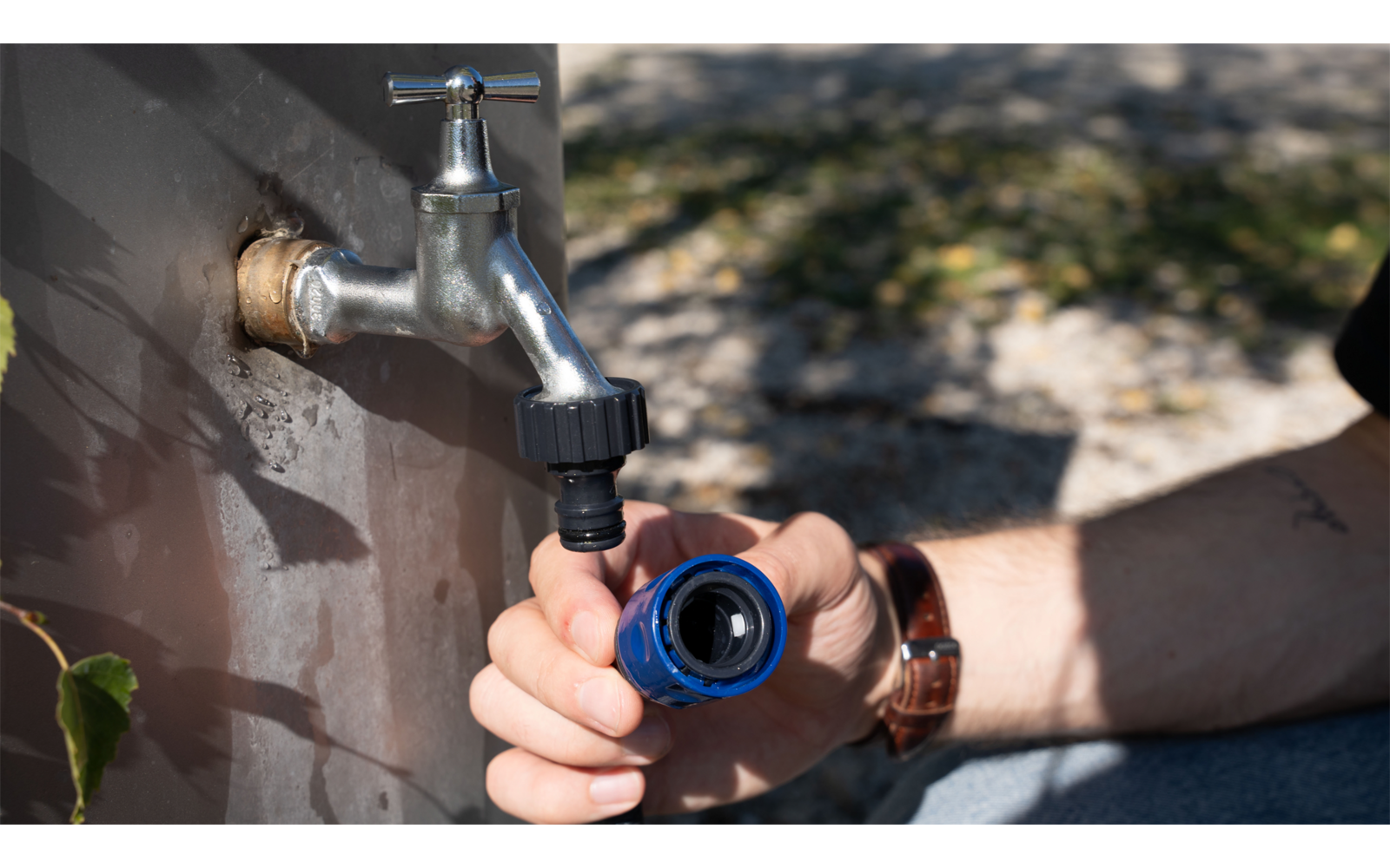 Metallwasserhahn an einer Außenwand, eine Hand hält einen blauen Schlauchanschluss, Unterarm mit Armbanduhr, Boden und Schatten im Hintergrund