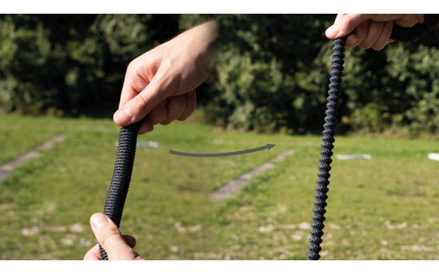 Zwei Hände halten jeweils ein schwarzes, geripptes flexibles Rohr, dazwischen ein gebogener Pfeil, im Hintergrund Rasen und Bäume
