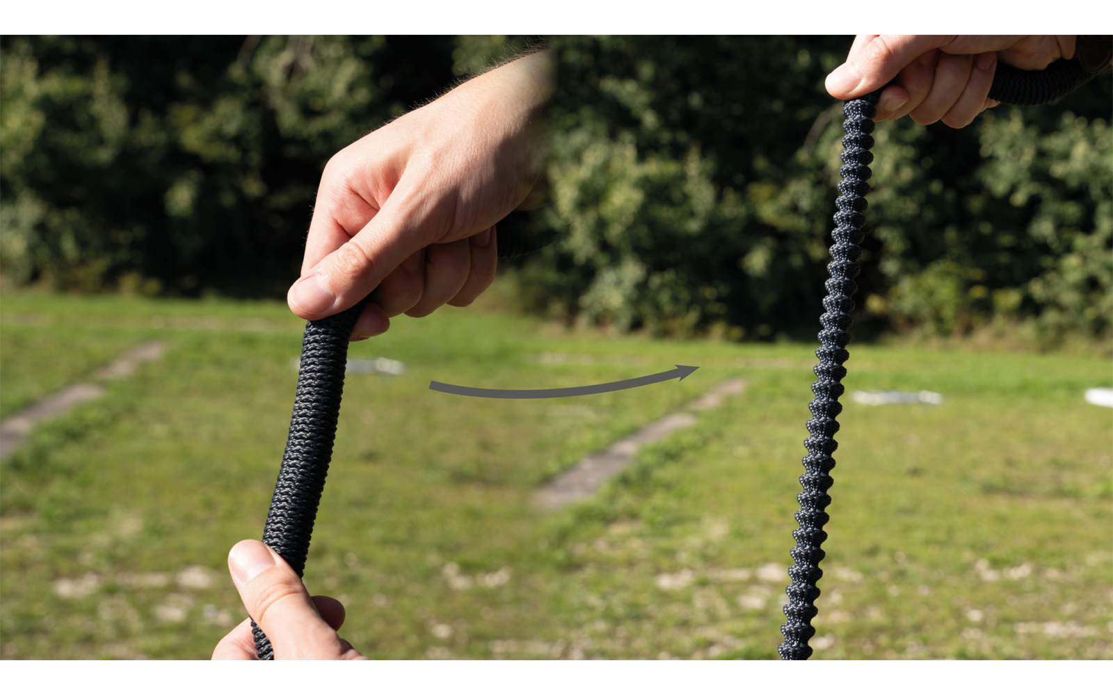 Zwei Hände halten jeweils ein schwarzes, geripptes flexibles Rohr, dazwischen ein gebogener Pfeil, im Hintergrund Rasen und Bäume