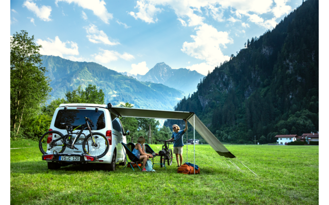 Camper bianco con tenda estesa su un prato davanti alle montagne, due persone vicino a un tavolo e sedie da campeggio, biciclette sul retro