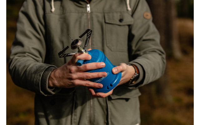 Brustbild einer Person in grüner Jacke, die mit beiden Händen einen kleinen Camping-Gaskocher an einer blauen Gaskartusche hält; verschwommener Wald im Hintergrund