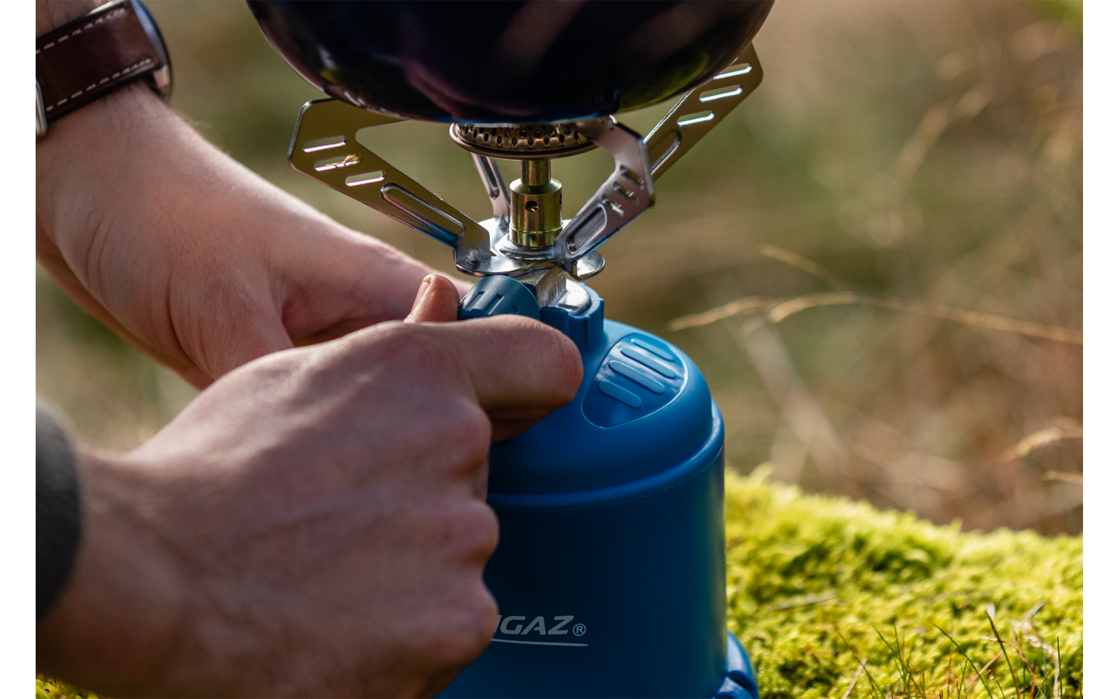Hände stellen einen Campingkocher auf einer blauen Gaskartusche ein, Topf auf dem Brenner, Moos und Gras im Hintergrund