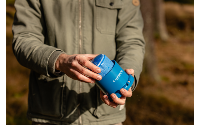 Oberkörper einer Person in grüner Jacke hält mit beiden Händen einen blauen, zylindrischen Trinkbehälter mit geöffnetem Deckel; unscharfer Waldhintergrund