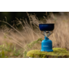 Blauer Camping-Gaskocher mit kleinem dunklem Topf auf moosbedecktem Stein, im Hintergrund hohes Gras