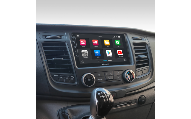 Innenansicht eines Fahrzeugs: manuelle Schalthebel im Vordergrund mit sichtbarem Schaltschema, mittig ein Touchscreen mit bunten App-Symbolen und Uhrzeit, darunter zwei Drehregler für Klima, links und rechts Luftausströmer