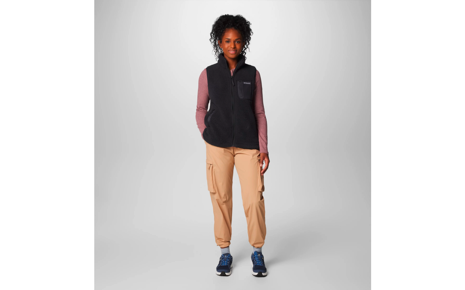 Person steht vor weißem Hintergrund, trägt schwarze Weste, rosa Langarmshirt, beige Hose und blaue Schuhe