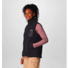 Frau trägt schwarzen ärmellosen Fleecepullover über rosa langärmligem Shirt vor grauem Hintergrund