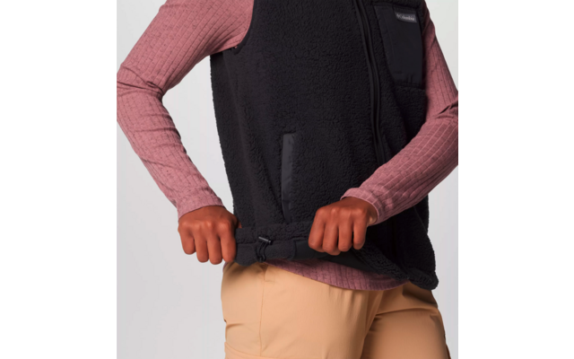 Person zieht einen schwarzen Reißverschluss an einer schwarzen Weste hoch, trägt rosa langärmliges Shirt und beige Hose