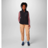 Person steht vor weißem Hintergrund, trägt schwarze Weste, rosa Langarmshirt, beige Hose und blaue Schuhe
