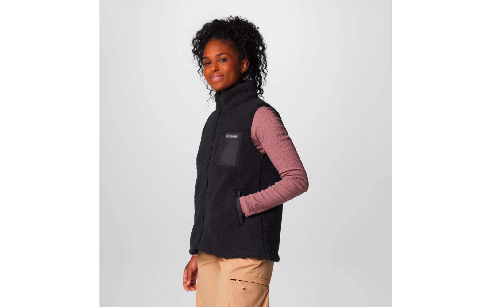 Frau trägt schwarzen ärmellosen Fleecepullover über rosa langärmligem Shirt vor grauem Hintergrund