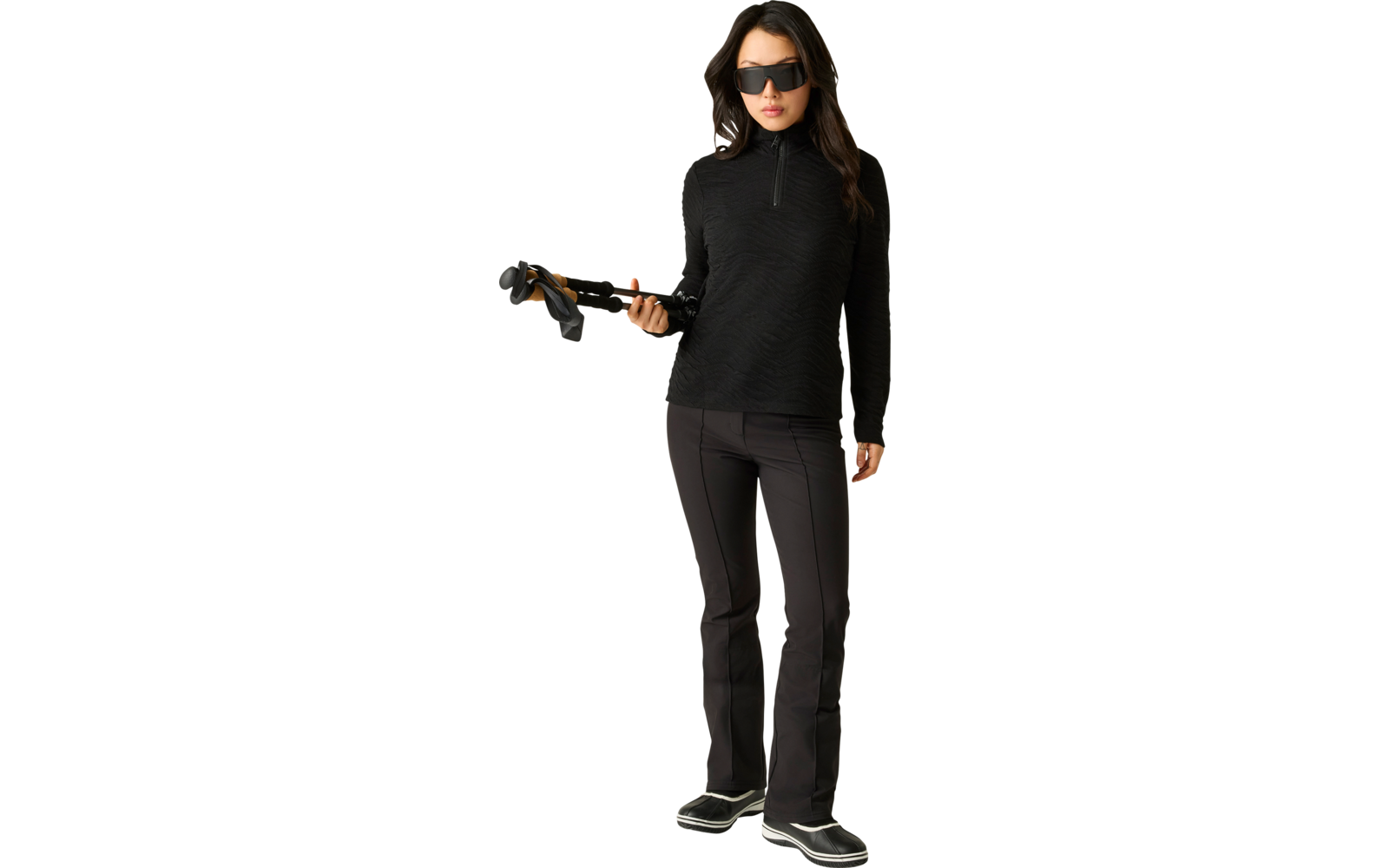 Person in schwarzem Langarmshirt mit kurzem Reißverschluss, schwarzer Hose und dunkler Sonnenbrille, steht vor hellem Hintergrund und hält Skistöcke in der rechten Hand