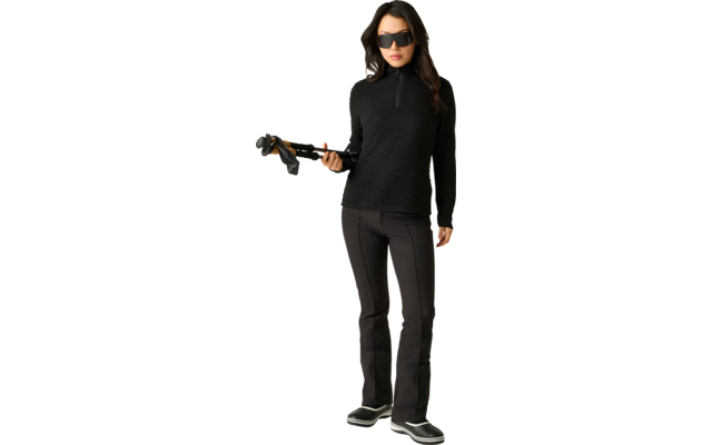 Person in schwarzem Langarmshirt mit kurzem Reißverschluss, schwarzer Hose und dunkler Sonnenbrille, steht vor hellem Hintergrund und hält Skistöcke in der rechten Hand