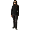 Person steht frontal, trägt schwarze Sonnenbrille, schwarzen Reißverschluss-Pullover mit langen Ärmeln, schwarze Hose und schwarze Schuhe vor hellem Hintergrund