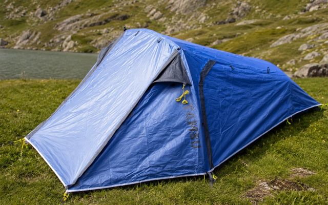 Tente de camping bleue sur l'herbe à côté d'un lac, collines rocheuses en arrière-plan