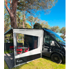 Furgoneta camper negra sobre césped junto a árboles; toldo gris-blanco acoplado con ventana transparente y sillas plegables rojas, cielo azul