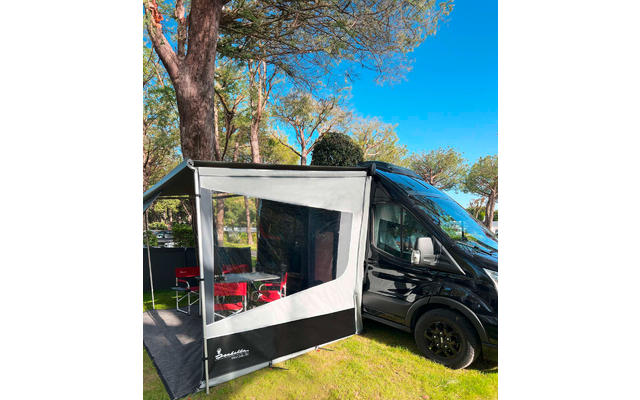 Furgoneta camper negra sobre césped junto a árboles; toldo gris-blanco acoplado con ventana transparente y sillas plegables rojas, cielo azul