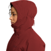 Persona de perfil mirando a la izquierda, rostro visible, lleva una chaqueta con capucha rojo oscuro, capucha levantada y cremallera cerrada