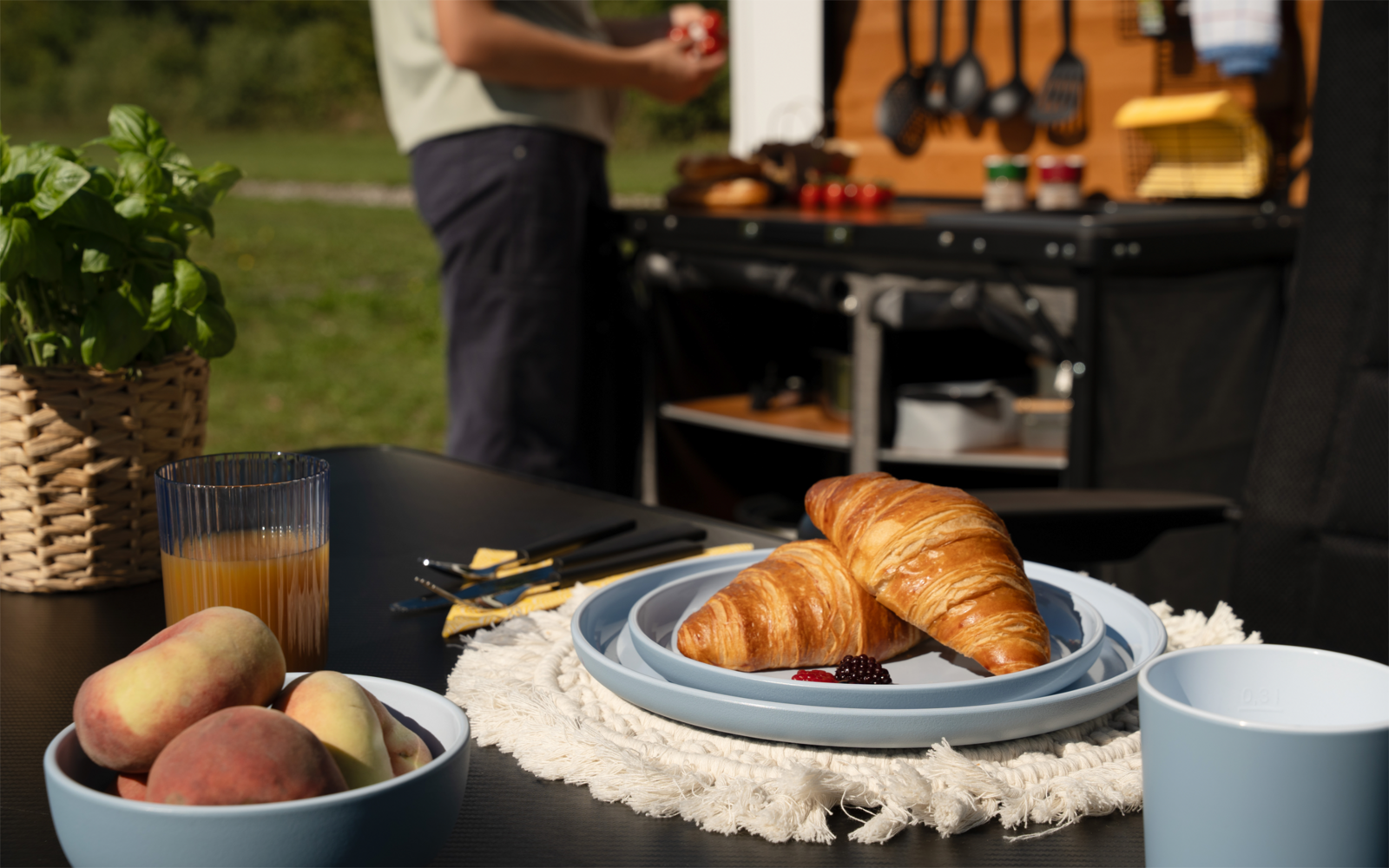 Zwei Croissants auf blauem Teller mit Marmelade, Schale mit Pfirsichen, Glas Saft, weiße Tasse, Pflanze im Korb, Person im Hintergrund vor einer Außenküchenstation