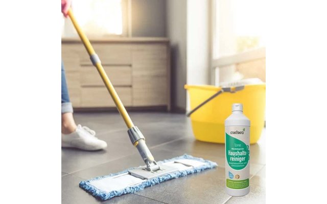 Füße in weißen Schuhen, ein Wischmopp mit blauem Pad auf dunklen Bodenfliesen, ein gelber Eimer und eine weiße Reinigungsflasche mit grünem Etikett Haushaltsreiniger im Vordergrund