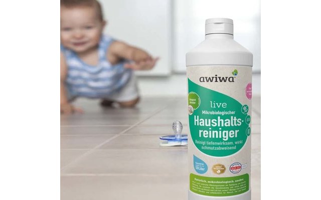 Flasche Haushaltsreiniger mit Aufschrift 'awiwa live' auf gefliestem Boden; Schnuller davor; unscharfes krabbelndes Baby im Hintergrund