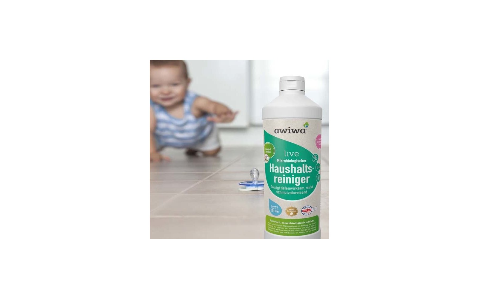 Flasche Haushaltsreiniger mit Aufschrift 'awiwa live' auf gefliestem Boden; Schnuller davor; unscharfes krabbelndes Baby im Hintergrund