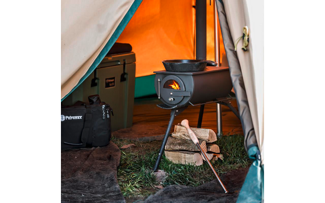Offenes Zelt mit kleinem Metallofen, sichtbare Flamme, Pfanne auf dem Ofen, gestapelte Holzscheite mit Axt, Kühlbox und Tasche am Eingang